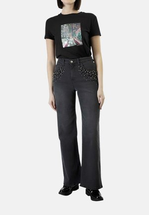 Persona che indossa una t-shirt nera con grafica e jeans larghi a vita alta scuri con dettagli borchiati sulle tasche anteriori e scarpe nere.