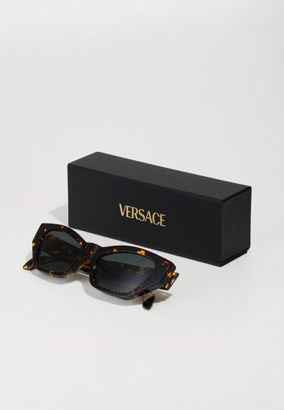Hörnchen-Sonnenbrille mit einem auffälligen Cat-Eye-Design. Dunkle Gläser und goldene Akzente. Wird in einer schwarzen Box mit "VERSACE"-Logo geliefert.
