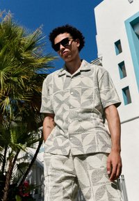 Koszula na krótki rękaw na guziki i dopasowane spodnie z teksturowanej tkaniny z jasnobeżowym i szarym wzorem liści, uzupełnione ciemnymi okularami przeciwsłonecznymi.