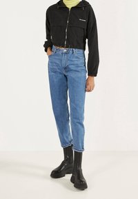 Black cropped jacket with zipper, two front pockets, paired with blue high-waisted jeans and black chunky ankle boots.