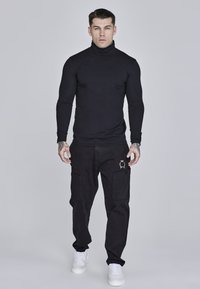 Schwarzer Rollkragenpullover, figurbetontes Design, kombiniert mit locker sitzenden schwarzen Cargo-Hosen mit Taschen und goldenen Stickereien, abgerundet durch weiße Sneakers.
