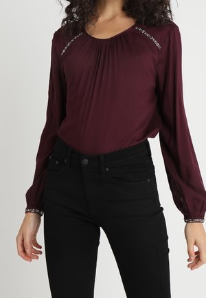 Femme portant un chemisier bordeaux à manches longues avec des ornements en strass aux épaules et aux poignets, associé à un pantalon noir taille haute.