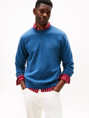 Felpa blu con collo a giro, indossata sopra una camicia a righe rosse. Il modello indossa pantaloni bianchi. Tessuto liscio, vestibilità casual e logo del brand visibile.