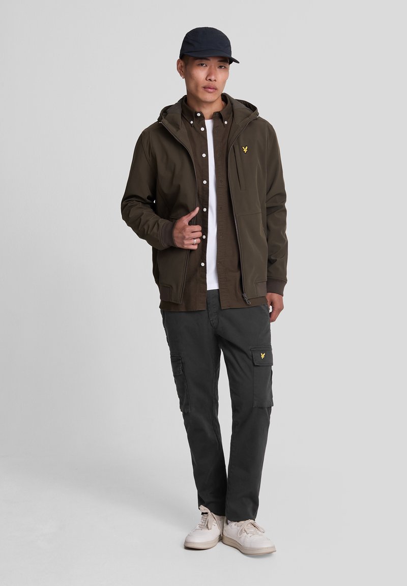 Dark olive green hooded jacket with a zip front, layered over a brown shirt and white t-shirt, paired with gray cargo pants and white sneakers.