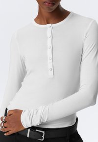 Witte lange mouwen shirt met een ronde halslijn en vijf knoopdetails aan de voorkant. Soepele textuur en een aansluitend ontwerp. Gepaard met een zwarte riem.