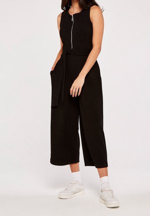 Kobieta ubrana w czarny kombinezon bez rękawów z suwakiem z przodu, szerokimi nogawkami, zawiązanym paskiem, białymi skarpetkami i białymi tenisówkami, stojąca na jednolitym tle.