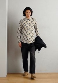 Langærmet, cremefarvet skjorte med et geometrisk mønster, parret med mørke jeans og brune sko. En sort jakke holdes i højre hånd.