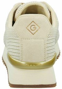 Beige Sportsschuh mit einem strukturierten Wildleder-Obermaterial, goldenen Akzenten an der Ferse und einer Gummisohle. Mit einem Logo auf der Rückseite.