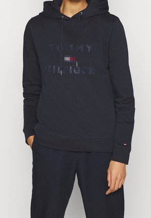 Osoba nosząca czarną bluzę z kapturem Tommy Hilfiger z troczkami i małym logo na lewej rękawie, połączoną z czarnymi spodniami.