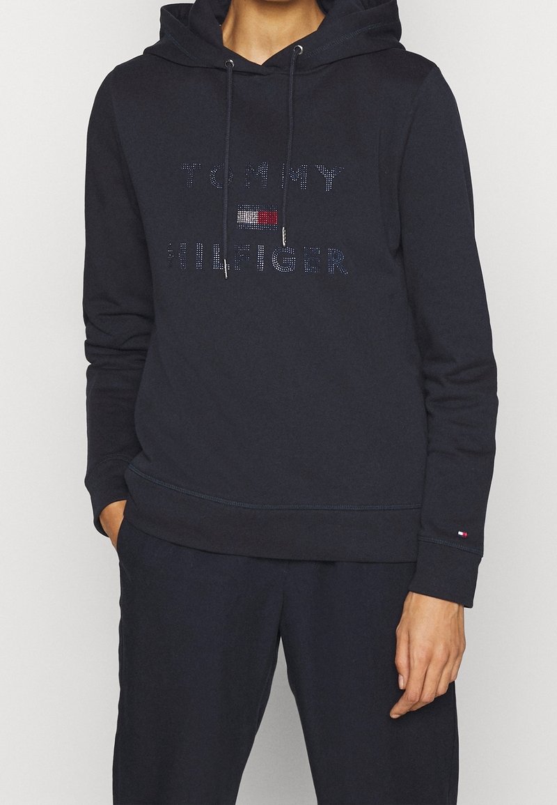 Personne portant un sweat à capuche noir Tommy Hilfiger avec cordons et un petit logo sur la manche gauche, associé à un pantalon noir.
