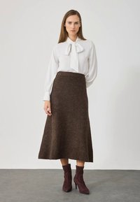 Femeie îmbrăcată cu o bluză albă cu mâneci lungi și fundiță, fustă midi maro și cizme cu toc la gleznă de culoare maro închis, stând pe un podea gri, în fața unui perete simplu.