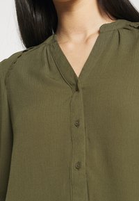 Blusa verde oliva con scollatura a V, chiusura con bottoni e tessuto testurizzato. Presenta delicati dettagli di cucitura lungo le spalle.