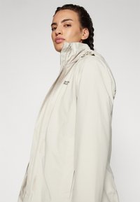 Giacca impermeabile beige con colletto alto, chiusura a zip e tasche frontali. Presenta una texture liscia e un discreto dettaglio del logo sul petto.