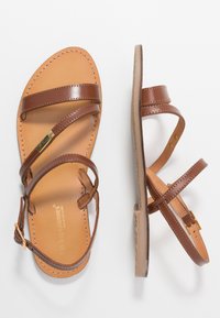 Sandalias de cuero marrón con dos correas finas y una hebilla en el tobillo. La plantilla es de un tono claro con costuras visibles, y la suela es plana.