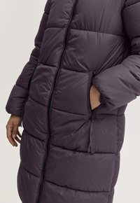 Kurtka puchowa w ciemnym fioletowym kolorze, z pikowanym wzorem, wysokim kołnierzem i kieszeniami na zamek. Tkanina o fakturze z gładkim wykończeniem.