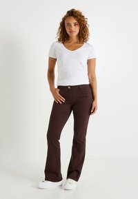 Jean flare marron avec fermeture à boutons, associé à un t-shirt blanc ajusté à col en V. Le modèle se tient avec les mains sur les hanches. Fond uni.