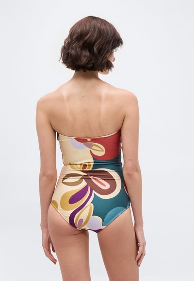 Γυναίκα με κοντά καστανά μαλλιά, που φοράει πολύχρωμο, στράπλες ολόσωμο μαγιό με αφηρημένα λουλουδένια σχέδια, στέκεται μπροστά σε απλό φωτεινό φόντο.