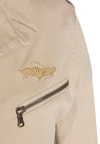 Veste légère beige avec col pointu, logo brodé doré et poche latérale avec fermeture éclair métallique. Texture lisse.