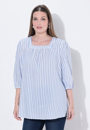 Kobieta o długich brązowych włosach, ubrana w niebiesko-białą bluzkę w pionowe paski z rękawami do łokcia i kwadratowym dekoltem, stojąca na białym tle.