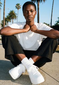 Giovane con capelli corti seduto a gambe incrociate sul marciapiede, indossa una maglietta bianca, pantaloni neri, scarpe da ginnastica bianche, con palme e cielo limpido sullo sfondo.