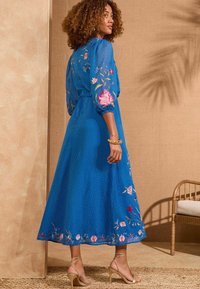 Blå knälång klänning med puffärmar, blommönster broderat på ärmar och fåll, texturerat tyg och en figursydd midja. Sandaler med remmar.