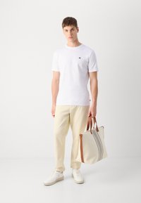 Hvid T-shirt med rund hals, beige casual bukser og hvide sneakers; holder en beige tote bag med brune håndtag og grå striber.