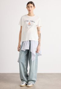 Cream t-shirt with printed text, layered over a blue striped button-up shirt, paired with wide-leg light blue jeans and silver sneakers.