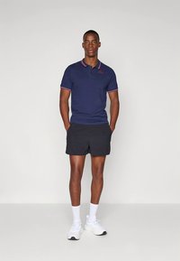 Tmavě modré polo tričko s červenými akcenty, v kombinaci s černými šortkami. Model má na sobě bílé tenisky a stojí před jednobarevným pozadím.