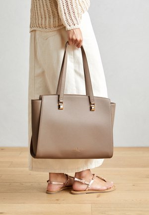 Brown synthetic tote bag with two long handles, square metallic accents, and a subtle logo. Worn with a light cream skirt and sandals.