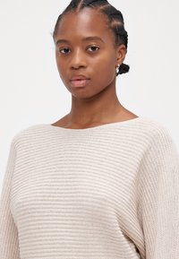 Μπεζ πλεκτό πουλόβερ με πλατιά λαιμόκοψη, μακριά μανίκια και απαλή, υφασμάτινη υφή. Το μοντέλο φοράει μικρές ασημένιες κρίκους.