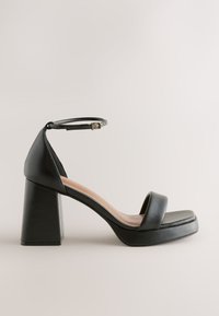 Czarna sandałka na wysokim obcasie z kwadratowym noskiem i szerokim paskiem z przodu. Posiada pasek wokół kostki z klamrą oraz masywny, prostokątny obcas.