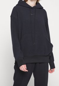 Námořnická fleecová mikina s klokaní kapsou, kapucí na šňůrku a pružnými manžetami. Na hrudi se nachází malé černé logo.