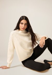 Kremowy sweter zdobiony kamieniami, o luźnym kroju, z dekoltem typu łódka i długimi rękawami, zestawiony z czarnymi obcisłymi spodniami.