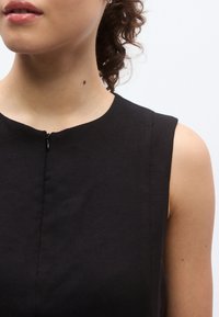 Μαύρο αμάνικο φόρεμα με στρογγυλή λαιμόκοψη, με πάνω ραφή και μικρή λεπτομέρεια φερμουάρ στο μπροστινό μέρος. Ομαλή υφή υφάσματος.