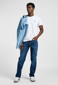 Camiseta blanca con el logo "Lee", chaqueta de mezclilla azul, pantalones vaqueros azul oscuro y zapatillas blancas con detalles en negro. Pose de pie frente a un fondo neutral.