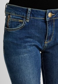 Jeans de denim azul oscuro con un corte ajustado, que cuentan con un bolsillo delantero con cremallera, costuras en contraste y un cierre de botón metálico.
