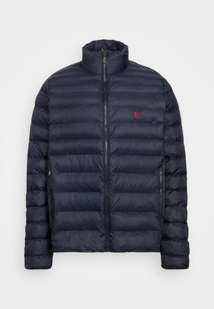 Giacca imbottita trapuntata blu navy con collo alto, cerniera frontale, tasche laterali con cerniera e piccolo logo ricamato rosso sul petto sinistro.