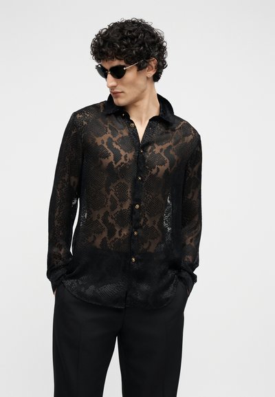 Homme portant une chemise noire transparente à motifs et boutons, un pantalon noir et des lunettes de soleil, les mains dans les poches, regardant sur le côté.