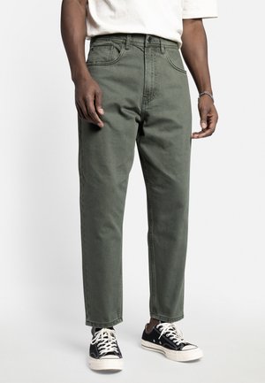 Mann trägt olivgrüne gerade geschnittene Hose, schwarz-weiße Sneakers und ein weißes T-Shirt und steht vor einem einfarbigen Hintergrund.