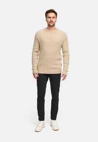 Vaahto beige neulepusero, jossa on kabelikuvio, yhdistettynä tummiin housuihin ja valkoisiin lenkkareihin. Malli seisoo neutraalia taustaa vasten.