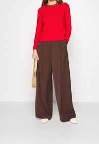 Punainen neulepaita pyöreällä kaula-aukolla, yhdistetty ruskeisiin leveälahkeisiin housuihin. Beige olkalaukku ja vaaleat kengät viimeistelevät asun.