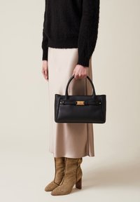Black leather handbag with a structured shape, gold hardware accent, and dual handles. Model wears a beige satin skirt and textured brown boots.