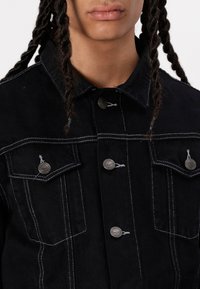 Schwarze Jeansjacke mit weißer Nähte, zwei Brusttaschen mit knöpfbaren Klappen und metallischen Hardware-Details. Strukturierter Stoff.