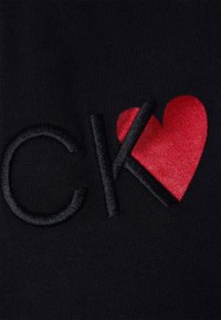 Czarna tkanina z haftowanym wzorem przedstawiającym litery "CK" w czerni oraz czerwone serce, widoczne szczegóły teksturowanego ściegu.