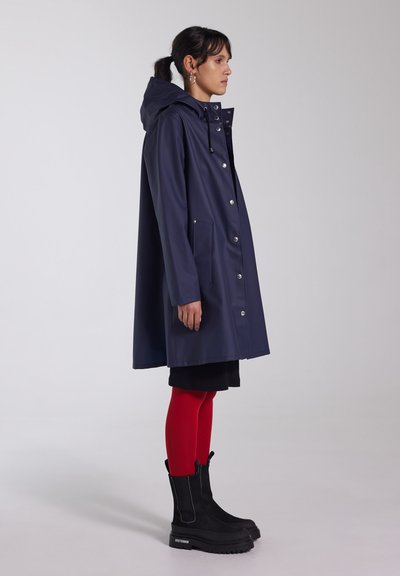 Marineblauwe waterdichte regenjas met een capuchon, voorzijde met drukknoopsluiting en zakken, gecombineerd met rode leggings en zwarte enkelboots met dikke zolen.