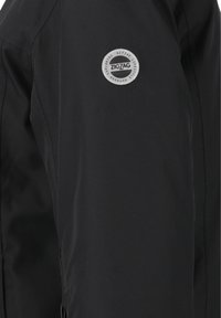 Schwarze Jacke mit weicher Textur, mit einem runden silbernen Logo auf dem linken Ärmel, das "ZIGZAG" und umgebenden Text zeigt.