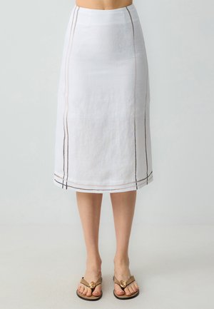 Jupe midi en lin blanc avec une silhouette droite, dotée de coutures brunes discrètes le long des côtés et de l'ourlet. Associée à des sandales simples.