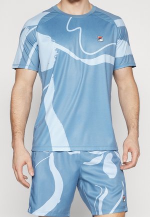 Homme portant un ensemble de sport bleu comprenant un t-shirt et un short avec des motifs abstraits clairs et de petits logos Fila sur la poitrine et la jambe.