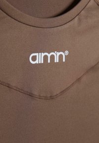 Pruun sportlik tank top, millel on ümmargune kaelus ja valge logo "aimn." Sile kangas, millel on pehme tekstuur.