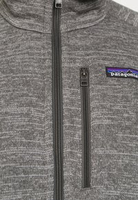 Veste en polaire grise avec un col haut, une fermeture éclair noire et un petit logo Patagonia avec un design de montagne coloré sur la poche poitrine.
